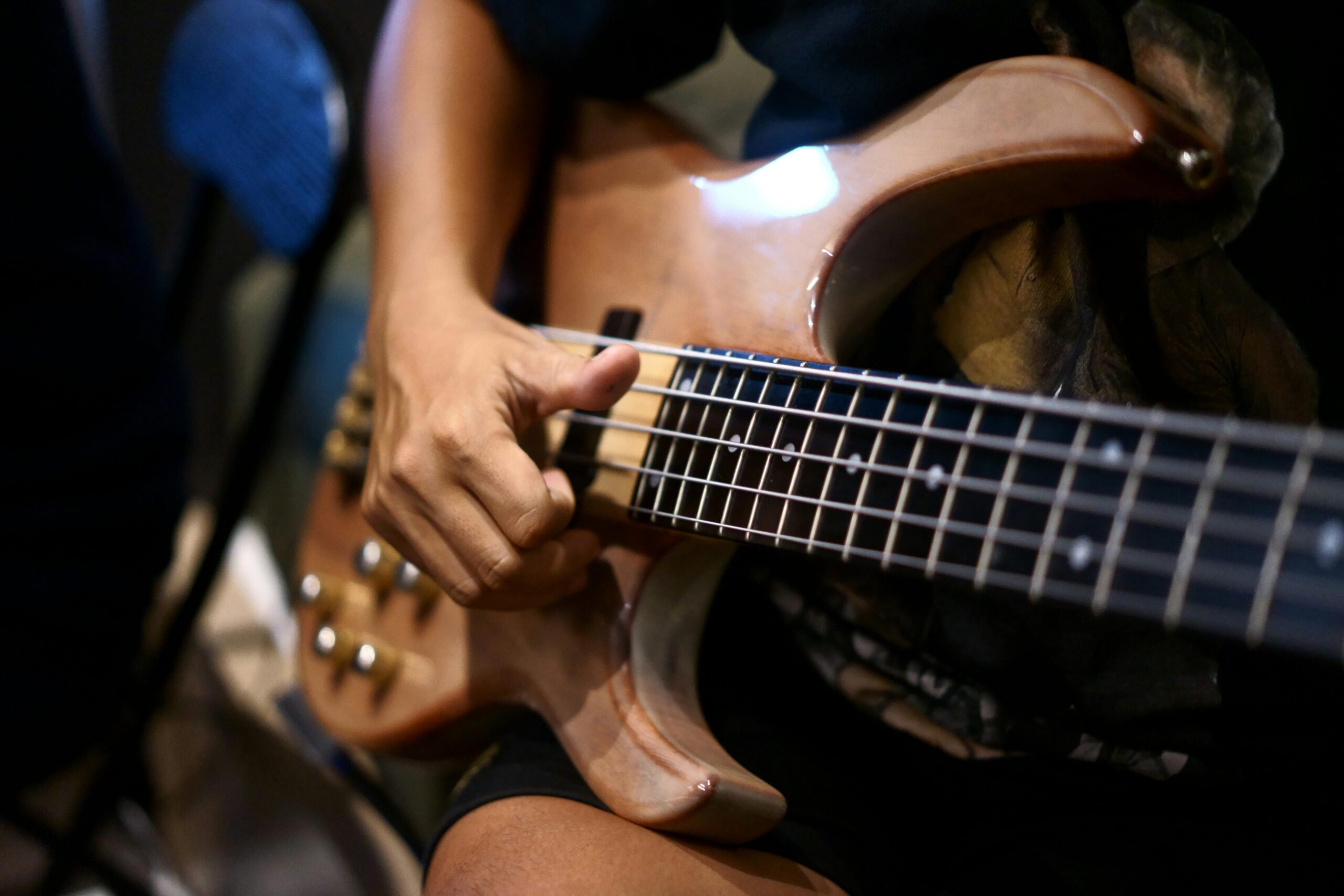 An adult playing a wooden electric bass guitar with focused detail on the strings and musician's hand.